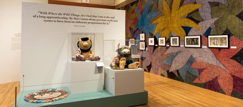 A wall covered in large, colorful flower illustrations and framed artworks appears on the right. On the left is a display of stuffed characters from the book Where the Wild Things Are and a wall quote by the artist Maurice Sendak.