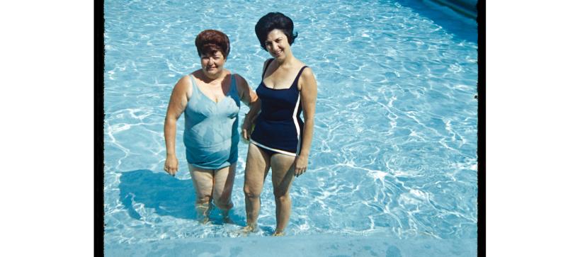 A mid-century photo taken of two women standing in a shallow swimming pool posing for the photo.