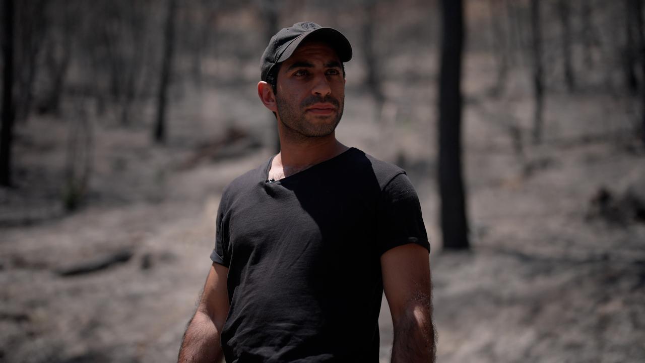 A man wearing a black cap and black t-shirt stands among burnt trees and stares into the distance