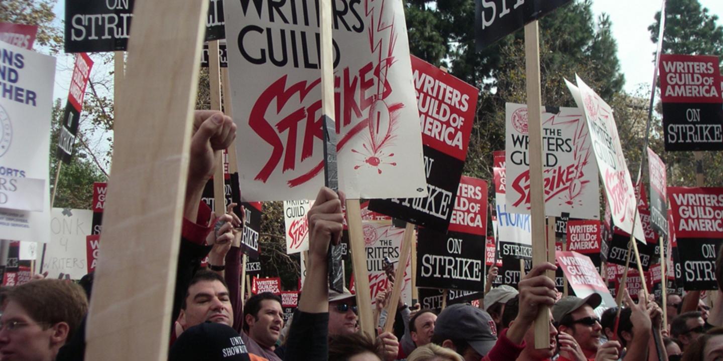 A crowd of people standing together holding signs that read Writers Guild of America On Strike