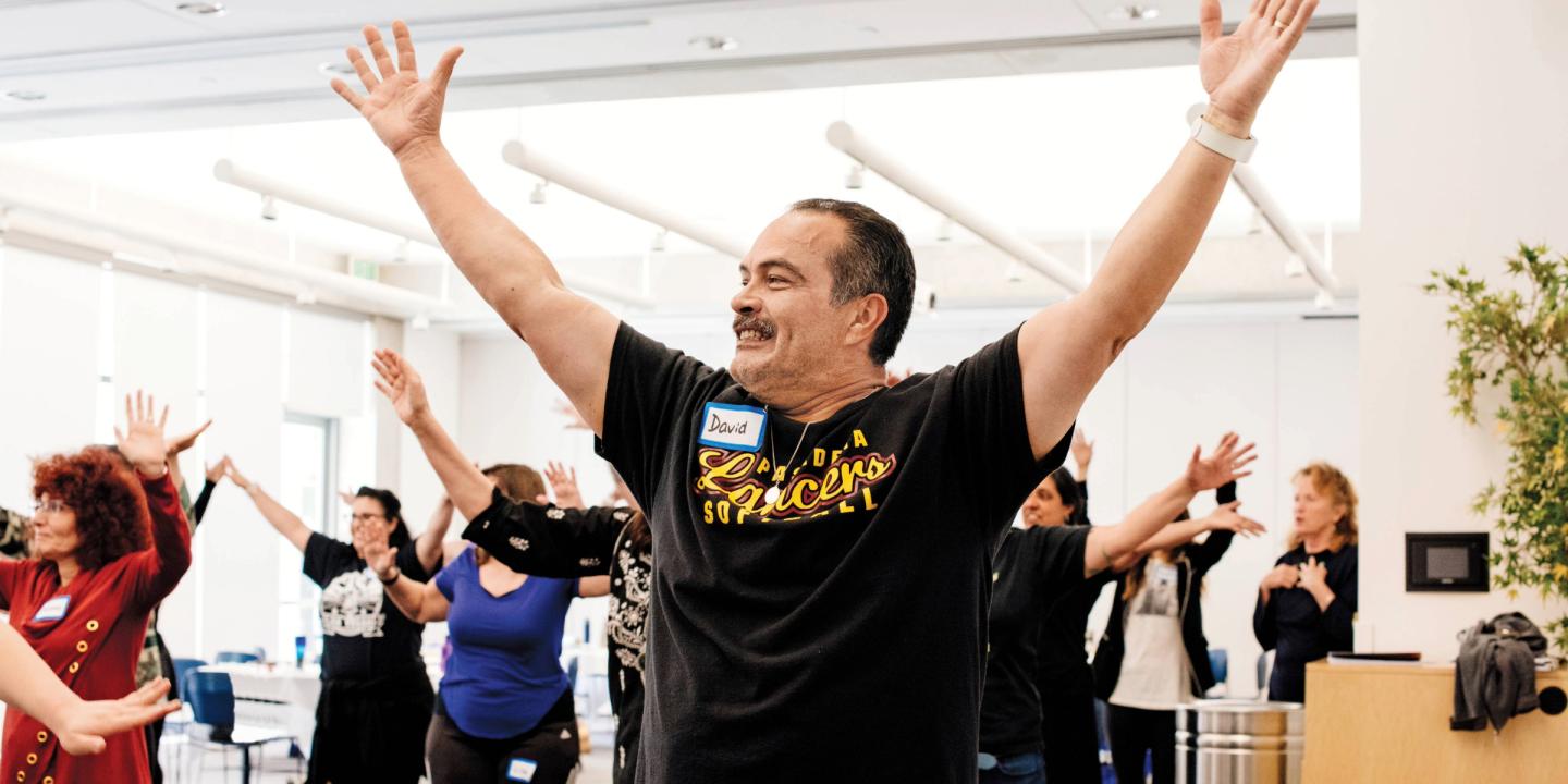 A group of people participate in a movement class at the Skirball. In the foreground is a man, smiling, and holding his hands above his head.