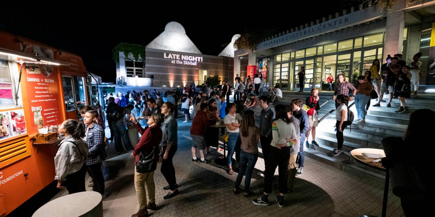 The front steps of the Skirball at night. People are standing around talking and ordering food from parked trucks. The words "Late Night!" are projected in lights on the side of the building.