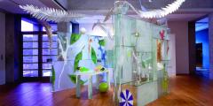 Photo of the Installation in a gallery space. Three glass walls surround a table with colorful objects. Above the walls, larger-then-life feathers suspend from the ceiling up into a skylight. 
