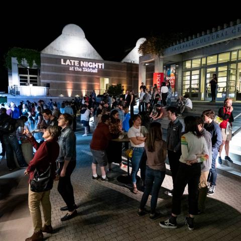 The front steps of the Skirball at night. People are standing around talking and ordering food from parked trucks. The words "Late Night!" are projected in lights on the side of the building.