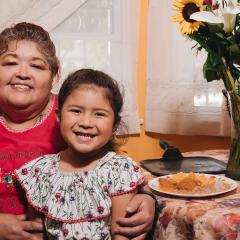 Photo of a woman and girl sitting together at a colorfully decorated table. They are both smiling at the camera. On the table are plates of food, a vase of colorful flowers, and a piñata.