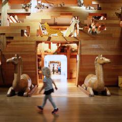A young girl walking through a floor to ceiling wooden ark with animals made from various materials.