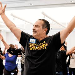 A group of people participate in a movement class at the Skirball. In the foreground is a man, smiling, and holding his hands above his head.
