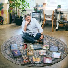 Dr. David Greenfield looking at the camera while sitting on a rug beside a collection of graphic novels and comic books on display.