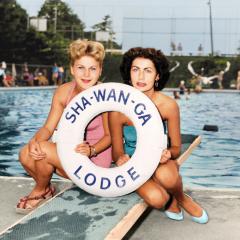 Two women pose in front of a swimming pool holding a life saver with the name Sha-Wan-Ga Lodge on it.