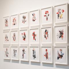 Photo of a gallery corner with a flat screen displaying a colorful video next to 18 framed collages hung in a grid.